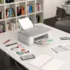 A Canon TS3451 printer is shown on a desk with books in the background, along with printed documents and a notebook. Produktbild-Vorschau
