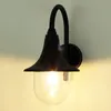 A black outdoor wall lamp with a clear glass shade is mounted on a wall. Náhled obrázku produktu