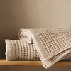 Two folded beige waffle-weave towels are stacked on a wooden shelf against a textured beige wall. Produktbild-Vorschau