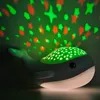 A whale-shaped night light projects stars and shapes onto a dark background. Produktbild-Vorschau