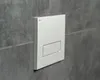 A close-up shot of a white toilet flush plate mounted on a gray tiled wall. Anteprima dell’immagine prodotto
