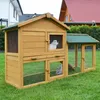 A wooden rabbit hutch with two levels and an attached run, situated on a green lawn in front of houses. Produktbild-Vorschau