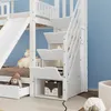 A white bunk bed with a slide and stairs with storage compartments, set against a light blue wall. Produktbild-Vorschau
