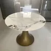 A round dining table with a marble-like ceramic top and a gold pedestal base, placed on a tiled floor in what appears to be an unfinished room. Produktbild-Vorschau