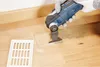 A person is using an oscillating multi-tool to cut into a wooden floor next to a vent. Produktbild-Vorschau