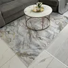 A rug with a marble abstract pattern in gray and gold is placed on the floor next to a sofa and a coffee table. Produktbild-Vorschau