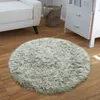 A round, fluffy gray rug is placed on a wooden floor in a living room setting, next to a white sofa and a white dresser. Produktbild-Vorschau