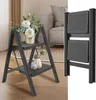 A dark gray, two-step folding ladder is shown in a living room setting, adorned with floral arrangements on its steps. A separate image of the ladder folded is shown on the right. Produktbild-Vorschau