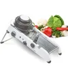 A white mandoline slicer with a gray food holder is shown in the foreground, with a variety of fresh vegetables in the background. Produktbild-Vorschau