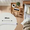 A bedside table with a digital clock, a cup of coffee, and a plant, next to a bed. Anteprima dell’immagine prodotto