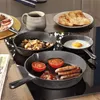 A breakfast scene with multiple cast iron pans on a stovetop, containing various breakfast items like sausages, eggs, tomatoes, and potatoes. Produktbild-Vorschau