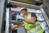 A construction worker wearing a hard hat, safety glasses, and a high-visibility vest is using a cordless drill to install a window frame. Produktbild-Vorschau