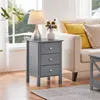 A grey nightstand with three drawers is placed next to a sofa with yellow and beige pillows. A lamp and a vase with flowers are on top of the nightstand. Produktbild-Vorschau