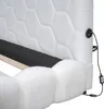 Close-up view of a white upholstered bed frame, showing the textured fabric and a power outlet with a cord. Náhľad obrázku produktu