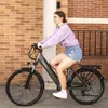 A young woman is riding an electric bicycle outdoors, with a brick wall and a pillar in the background. Produktbild-Vorschau