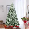 A flocked artificial Christmas tree decorated with snow, standing in a room with gifts at its base and a fireplace with stockings. Produktbild-Vorschau