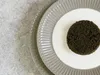A close-up overhead view of a serving of black caviar on a white plate, which is placed on a grey ribbed plate, all set against a textured grey surface. Produktbild-Vorschau