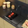A modern kitchen sink with a black granite sink, a black faucet, and tomatoes being washed under the running water. There are also jars of nuts and wooden bowls on the countertop. Produktbild-Vorschau