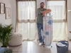 A man is leaning on an ironing board in a room with curtains and a plant. Produktbild-Vorschau