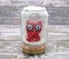 A decorative snow globe with a red owl inside, placed on a wooden surface with scattered artificial snow. Voir les images du produit