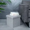 A gray fabric storage ottoman sits on a tiled floor next to a gray sofa, with a plant in the background. Náhled obrázku produktu