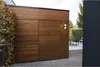 A modern wooden garage or shed with a built-in door, set in a garden with gravel and greenery. Podgląd zdjęcia produktu