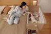 A woman is lying on a bed next to a baby bassinet, gently touching the baby inside. Produktbild-Vorschau