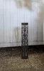 A black, hexagonal solar pathway light with a decorative cut-out pattern stands on a gravel surface in front of a white corrugated wall. Produktbild-Vorschau