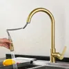 A hand is holding a pull-down kitchen faucet spray head, with water spraying into a sink. A yellow fruit, possibly a lemon or mango, is in the sink. Produktbild-Vorschau
