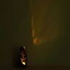 A dark, oval-shaped solar lantern with a tree silhouette cut-out is placed on a surface, casting a warm yellow light and shadow pattern on a dark wall. Produktbild-Vorschau