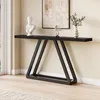 A black console table with a triangular base is placed against a cream-colored wall, near a window. Decorative items, including a bust and a vase with greenery, are displayed on the table. Produktbild-Vorschau