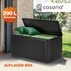 An open black storage box with green cushions and pillows inside, placed on a patio with a stone wall and trees in the background. Produktbild-Vorschau