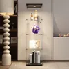 A well-lit display cabinet containing various items, including bottles, a figurine, books, and decorative objects, situated in a modern interior. A large, textured lamp is visible on the left. Náhled obrázku produktu