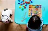 A child playing with a wooden bead toy on a blue table, with a French bulldog lying on the floor nearby. Produktbild-Vorschau