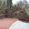 A rustic stone wall with a decorative arched window opening, set in a garden with brick and gravel flooring. Produktbild-Vorschau