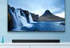 A black soundbar is placed on a white cabinet below a large television displaying a scenic waterfall and mountain landscape. Voir les images du produit