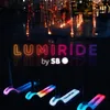 Four illuminated scooters are lined up in the foreground, with a neon sign reading "LUMIRIDE by SB" above them. In the background, a fountain with colorful lights is visible. Produktbild-Vorschau