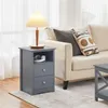 A dark gray nightstand with two drawers and an open shelf sits next to a beige sofa with decorative pillows. A lamp with a pleated shade is on top of the nightstand. Produktbild-Vorschau