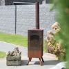 A rusty metal outdoor fireplace with a chimney stands on a paved patio next to a wooden crate filled with firewood. A stone fence and green grass are in the background, with blurred foliage in the foreground. Produktbild-Vorschau