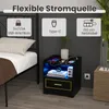 A black nightstand with LED lighting, featuring USB and Type-C charging ports, is shown in a bedroom setting next to a bed. The nightstand has a drawer and is displaying electronic devices. Náhled obrázku produktu