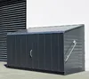 A dark gray metal storage box with a slanted roof, positioned outdoors on a paved surface next to a white wall and a building with dark horizontal blinds. Náhľad obrázku produktu