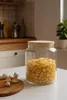A glass storage jar filled with pasta sits on a white table, with garlic and a wooden cutting board nearby. The background shows a kitchen counter with shelves and appliances. Produktbild-Vorschau