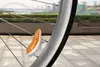 A close-up of a bicycle wheel with a small, orange and white hamster-shaped accessory attached to the spokes. The background shows a blurred cityscape with a bridge and trees, suggesting the bike is in motion. Podgląd zdjęcia produktu