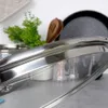 A close-up shot of a glass pot lid with a stainless steel rim, resting on a countertop. Other kitchen items like a frying pan and a plant are visible in the background. Voir les images du produit