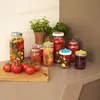A collection of glass jars filled with preserved food items, such as tomatoes, peppers, and other vegetables, arranged on a kitchen countertop next to a potted herb plant. Produktbild-Vorschau