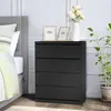 A black dresser with four drawers is placed next to a bed in a bedroom. There are two photo frames and a vase with flowers on top of the dresser. A plant is also visible to the right of the dresser. Voir les images du produit