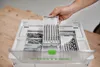 A hand is shown placing a set of drill bits into a white Festool Systainer³ tool case. Voir les images du produit