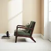 A green armchair with a small pillow sits on a white floor against a cream-colored wall with soft shadows cast by sunlight. Produktbild-Vorschau