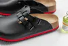 A close-up shot of a pair of black Birkenstock clogs with red trim, placed on a light gray surface. A small bottle of yellow liquid is visible in the background. Anteprima dell’immagine prodotto