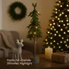 A small LED-decorated Christmas tree stands on a table next to a white reindeer figurine, a lit candle, and a gift-wrapped box. A larger, out-of-focus Christmas tree with lights is in the background, along with a wreath on the wall. Produktbild-Vorschau